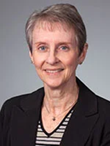 An image of Dr. Terry Jones. She has short gray hair, pale skin, and is smiling. She wears a black coat, striped shirt, and a necklace. The image is taken in front of a gray background.