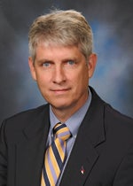 An image of Dr. David Collier. He has tanned skin and gray-brown hair. He is wearing a suit, as well as a tie with blue and yellow stripes.