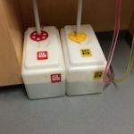 A picture of two small white chemical waste tanks sitting on a gray floor. The left tank has a red lid and accents, whereas the right has a yellow lid and accents. Each tank has a pipe leading into the top, and they sit directly next to each other tucked into a nook.