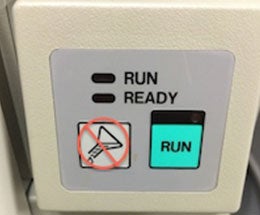 An image of the side of a machine. It has lights next to two printed words- 'RUN' and 'READY'- whcih indicate if the machine is currently turned on or if it is ready for activation. Below the indicator lights, there is a blue button which says 'RUN', which is used to start the machine. To the left of the blue button, a symbol indicates that chemicals are not to be poured into the machine.