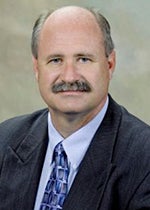 An image of Dr. Robert Lust. He has pale skin, gray hair, and a moustache. He wears a black suit and a blue patterned tie.