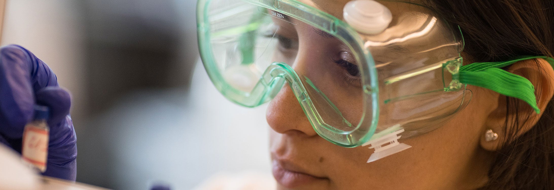 A close-up image of a scietist with green safety googles picking up a vial.