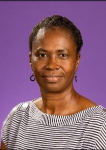 An image of Ms. Vickie Staton. She has short and curly brown hair, as well as dark skin. She is wearing earrings and a black-and-white striped shirt. Her picture was taken in front of a purple background.