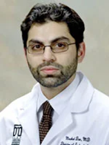 An image of Dr. Moahad Dar. He has black hair and facial hair, and pale skin. He is wearing glasses, a tie, and a lab coat.