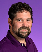 An image of Dr. Zachary Domire. He has dark brown hair, a beard, and tanned skin. He is wearing a dark purple shirt, and the image was taken in front of a lighter purple background.