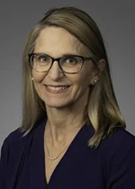 An image of Dr. Suzanne Lazorick. She has long blonde hair, pale skin, and is smiling. She is wearing glasses, earrings, and a necklace. The image is overlaid on a gray background.
