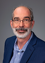 An image of Dr. Jaques Robidoux. He has mid-tone skin, gray hair, and a beard. He is wearing a blue suit and a blue striped shirt. He also has glasses.