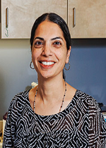 An image of Dr. Shahnaz Aziz. She has tanned skin and black hair tyed back in a ponytail. She wears a necklace, earrings, and a black shirt with white swirls. She is smiling brightly.