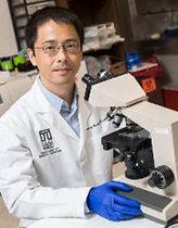 An image of Dr. Li Yang. He has tanned skin and black hair, and wears glasses. He is wearing a Brody School of Medicine lab coat and is shown in the lab in front of a microscope.