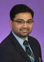 An image of Dr. Shivajirao Patil. He has black hair, facial hair, and light brown skin. He has glasses, and is wearing a black suit and a black patterened tie. His image is taken in front of a purple background.