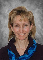 An image of Dr. Suzanne Lea. She has medium-length blonde hair, tanned skin, and is smiling brightly. She is wearing a blue scarf and a black blouse. She stands in front of a patterened gray background.